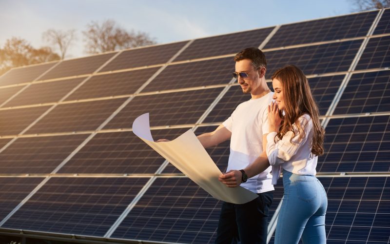 Young beautiful couple on a sunny day is standing by a solar module and looking into a big white paper plan, thinking about future, concept of bright future